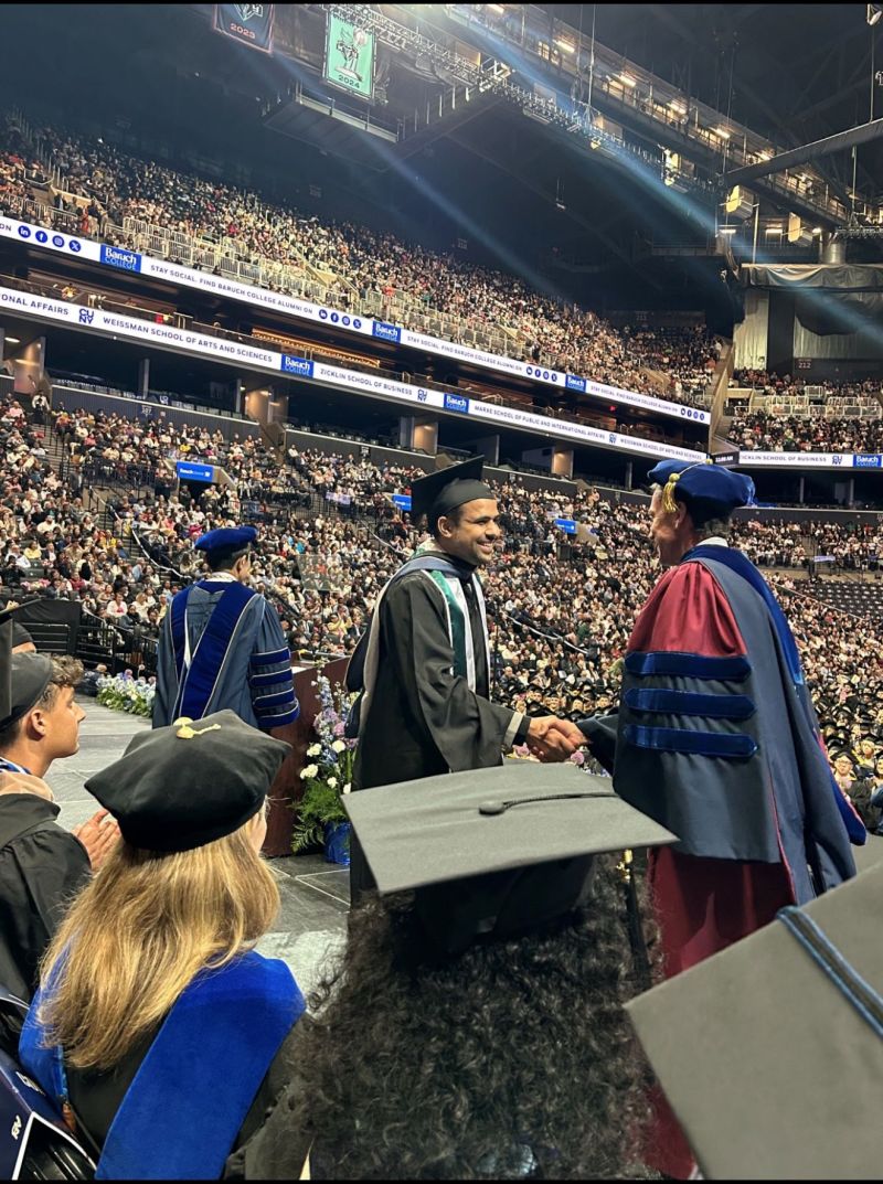 Salman in graduation regalia, shaking hands on stage at his MBA commencement, with the arena audience behind him.